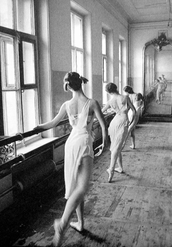 Black-and-white photo of three ballerina's lined up at a bar set against a wall of windows away from the lens, with their backs turned toward the camera in the midst of a ballet exercise, their right leg extended back.