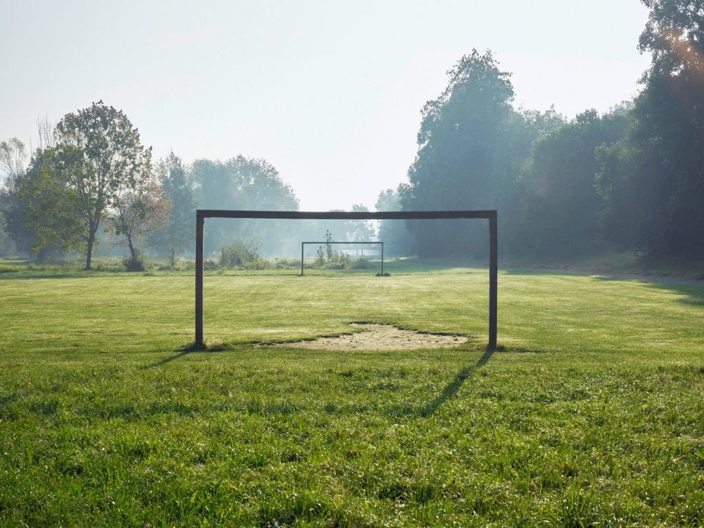A photo of a grassy field lined by trees and low hanging fog with two metal soccer goals without their nets.