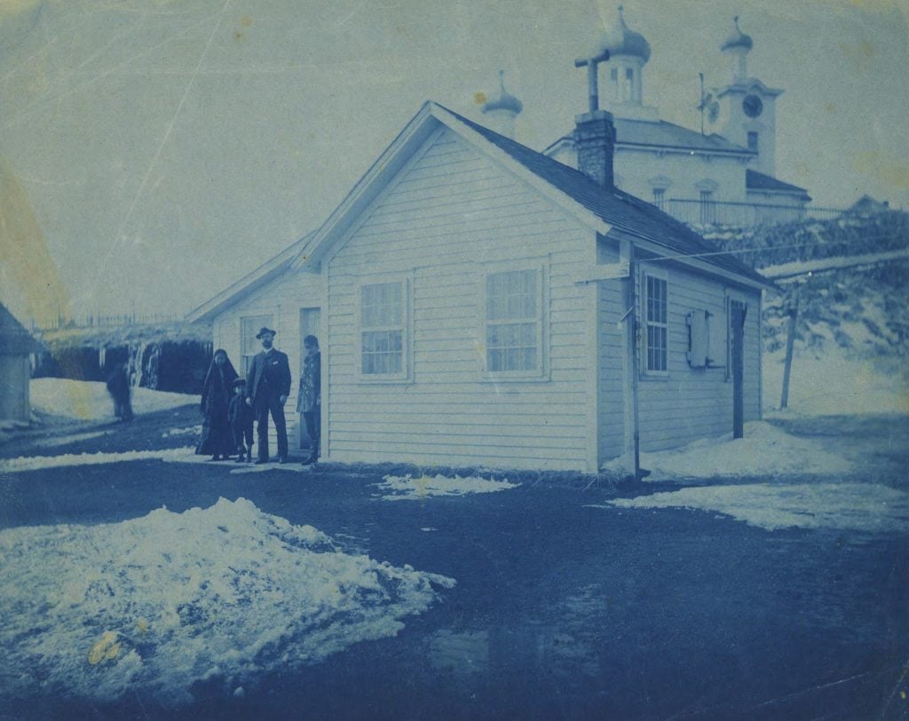 Cyanotype of two men, woman, and boy standing beside a wood-framed house at the end of a driveway