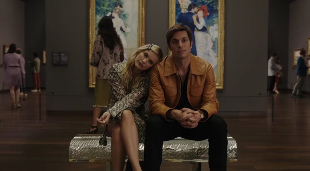 A blonde woman rests her head on the shoulders of a handsome man in a tan leather jacket as they sit on a bench in front of two Renoir paintings of life-sized couples dancing at the Musée d'Orsay in this scene from Emily in Paris.