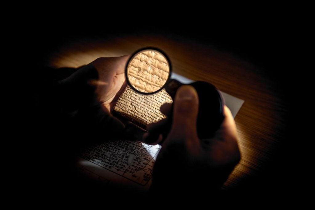 A darkened photograph of a pair of hands examining a cuneiform tablet with a magnifying glass.