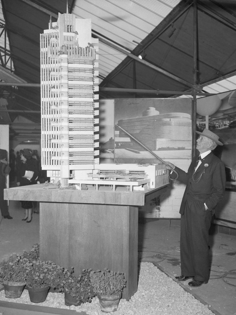 A man in a suit and hat, architect Frank Lloyd Wright, pointing a walking cane at a model of a skyscraper