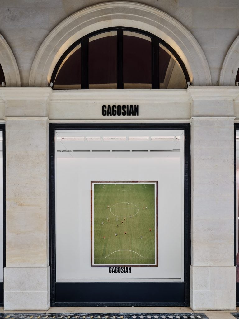 A photograph of the exterior of Gagosian gallery in Paris, with a lit display window featuring a green photograph of a soccer field from their "Art of the Olympics" show