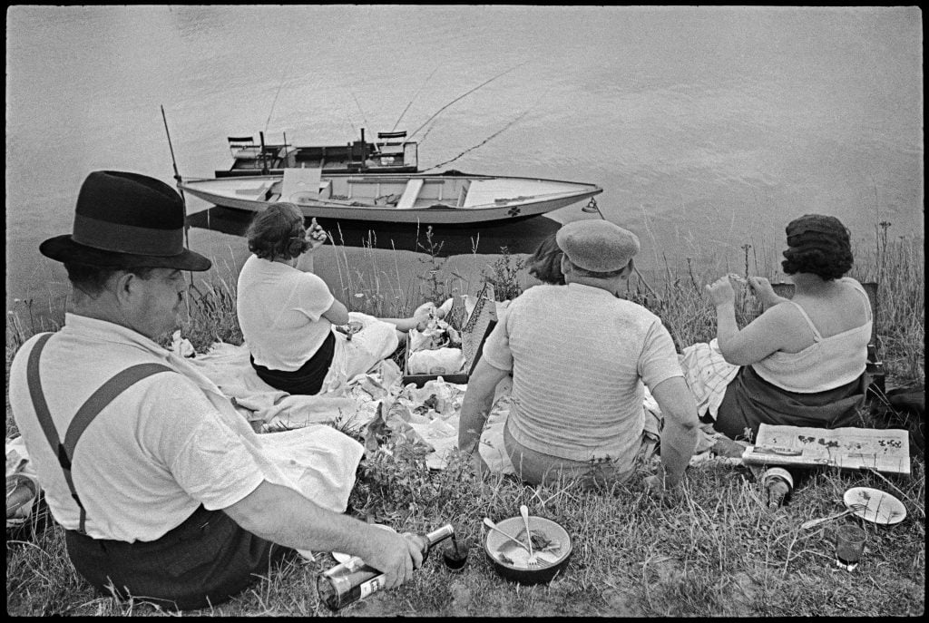 Black-and-white photo of four individuals sitting facing away from the viewer on a grassy edge of a body of water surrounded by picnic items. A small vacant boat is in the water. Included in the book the Power of Photography produced by Peter Fetterman Gallery.