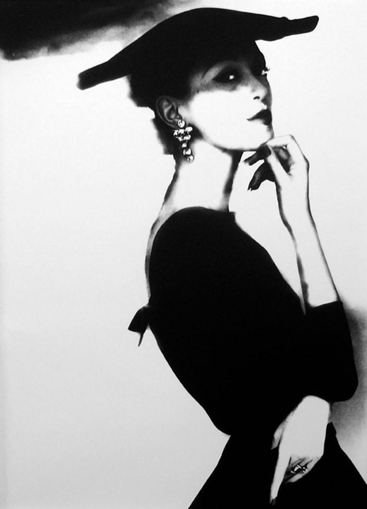 A back-and-white half-portait of a woman wearing a black frock and flat black hat in profile looking over at the camera with her left arm crossed and right hand up under her chin.