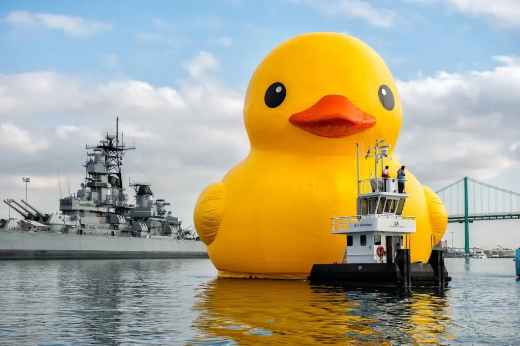 Mama Duck, the "World's Largest Rubber Duck," a giant inflatable yellow rubber duck towers in the water above the the USS Iowa battleship.