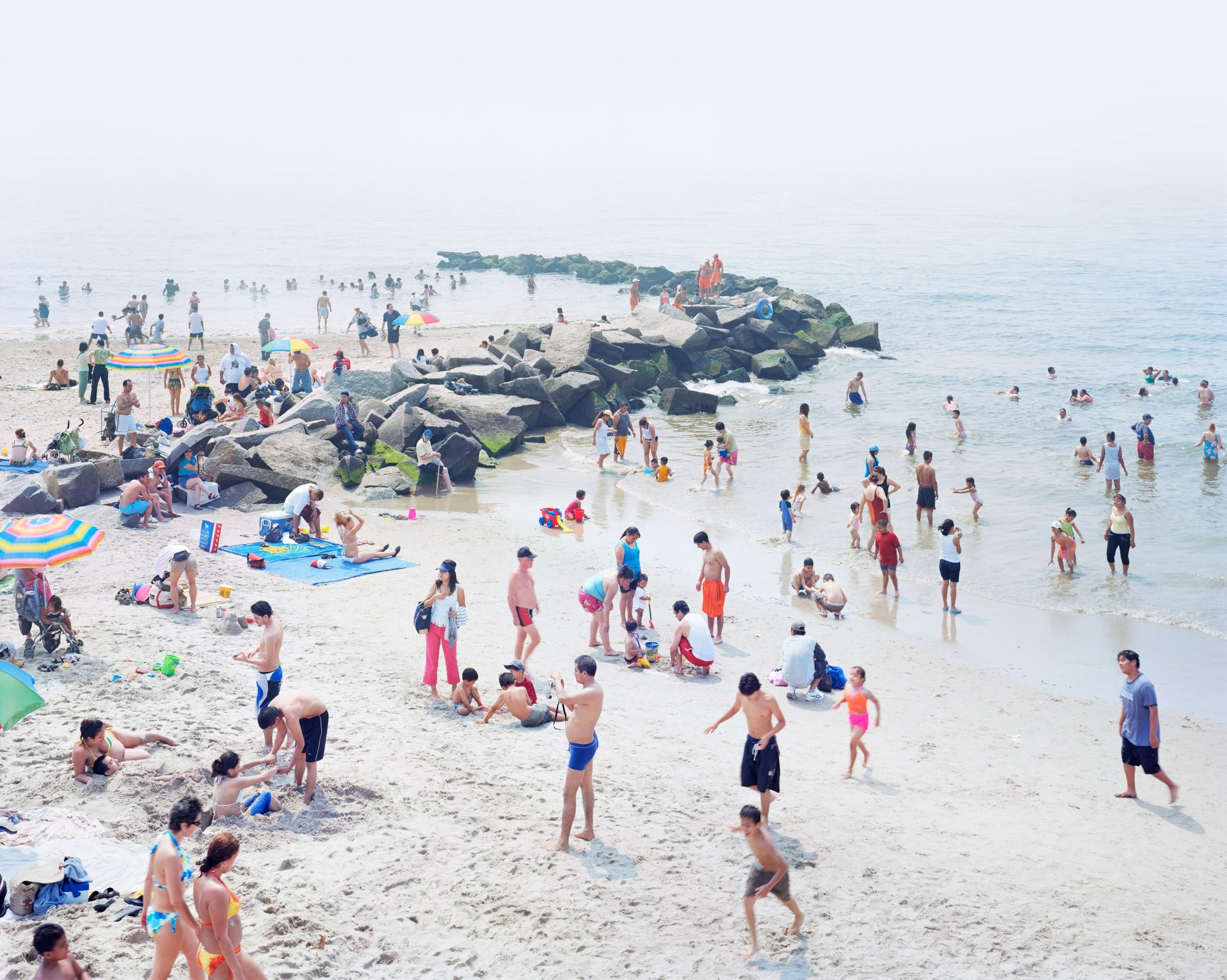 Summer beach scene photography by Massimo Vitali showing dozens of people in the sand and in the water wearing colorful bathing suits, very bright but the horizon is obscured by white fog.