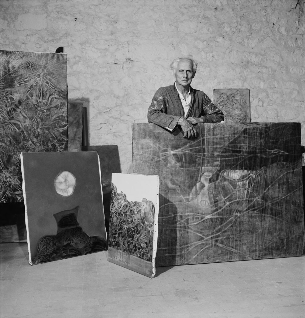 An elderly man, artist Max Ernst, standing with a group of his paintings