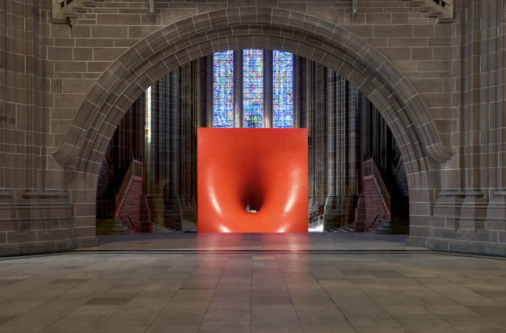 A photograph of a large sculpture of a red cube with an entrance hole in its front by Anish Kapoor installed in front of a window in the stony Liverpool Cathedral