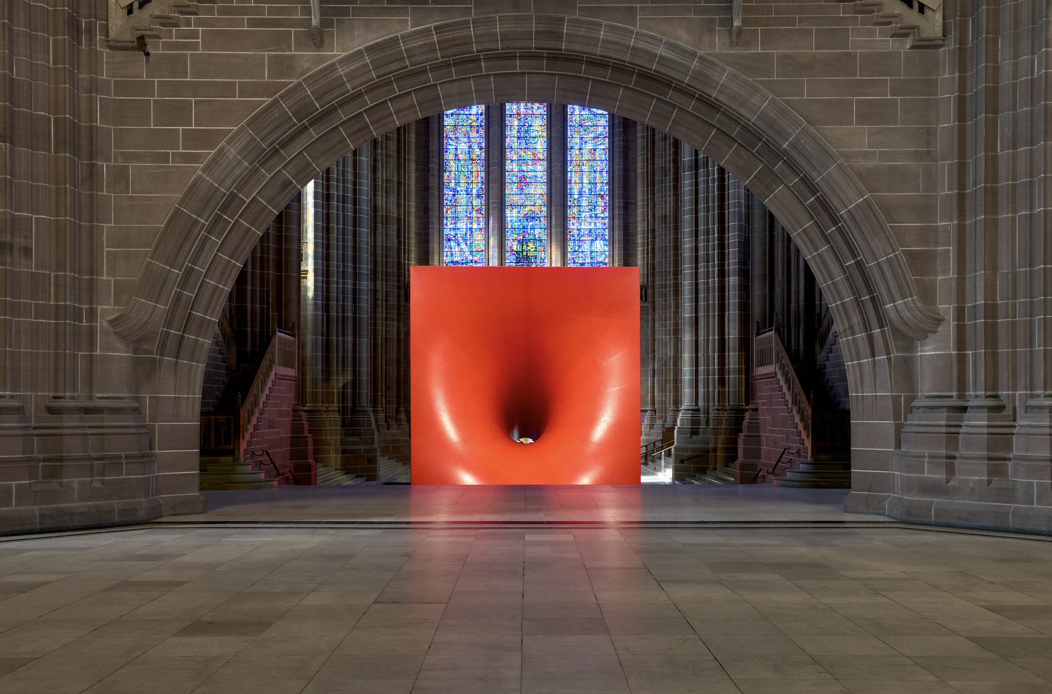 A photograph of a large sculpture of a red cube with an entrance hole in its front by Anish Kapoor installed in front of a window in the stony Liverpool Cathedral