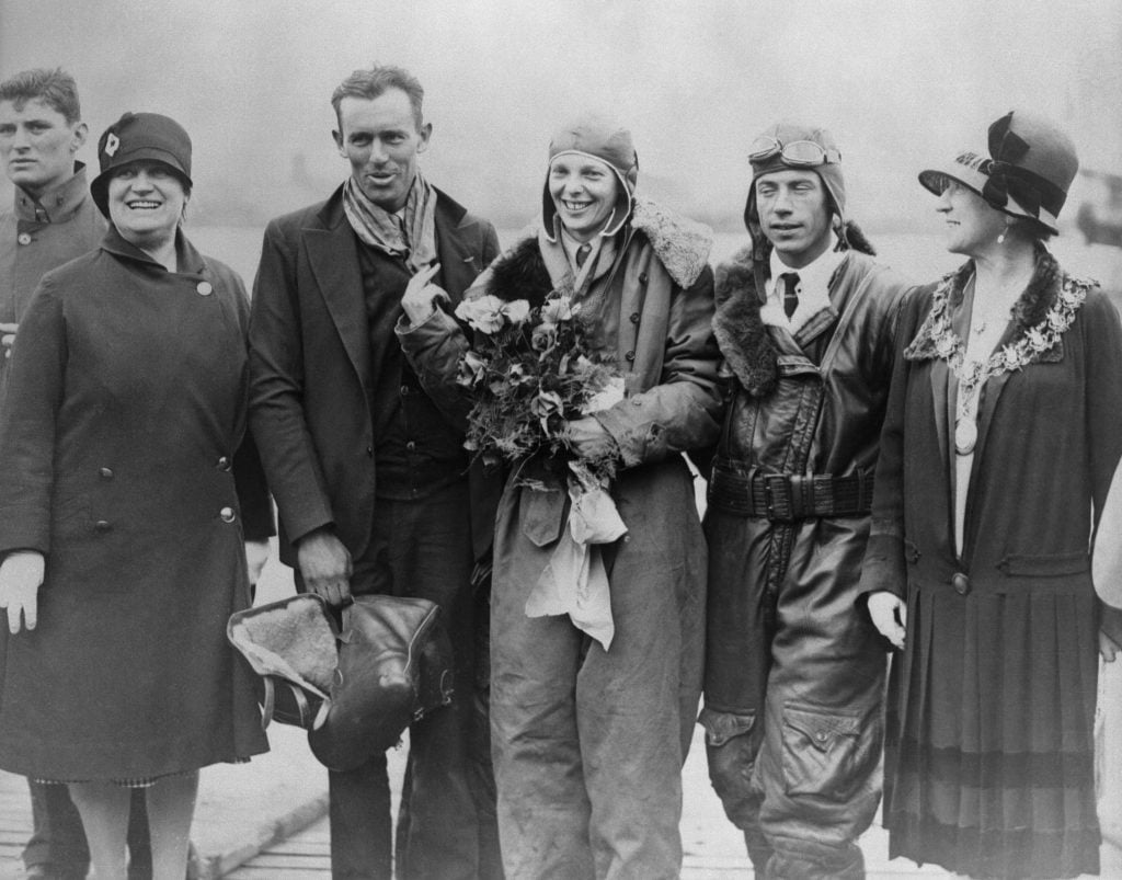 A black-and-white photo showing aviator Amelia Earhart holding flowers and flanked by four other people