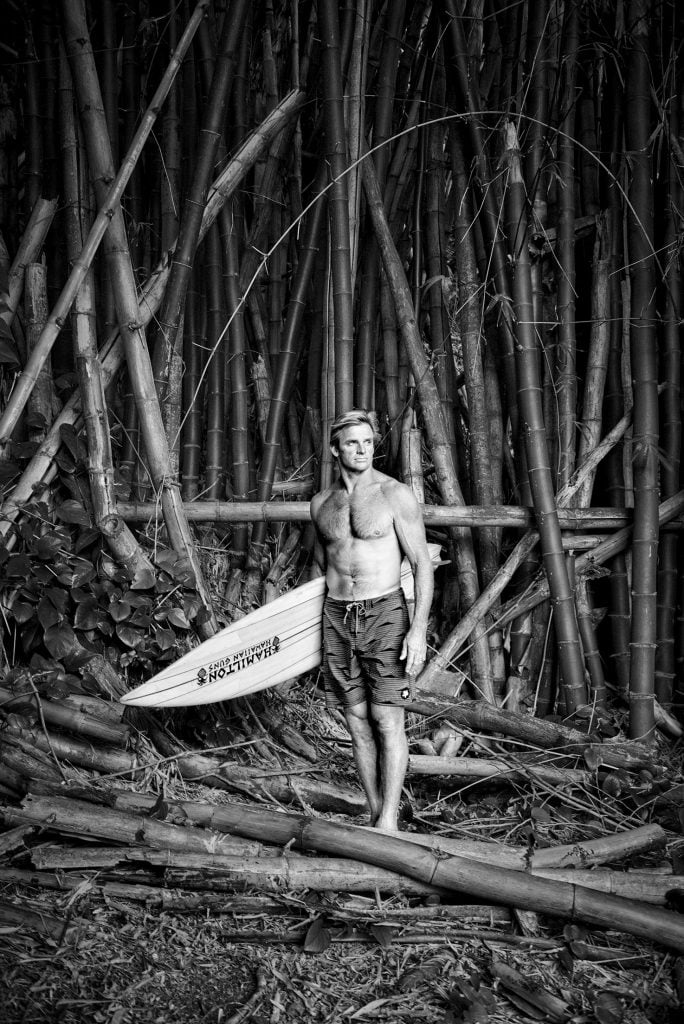 Black and white portrait of Olympics medalist Laird Hamilton wearing swim trunks and holding a surfboard standing in front of a copse of bamboo trees.