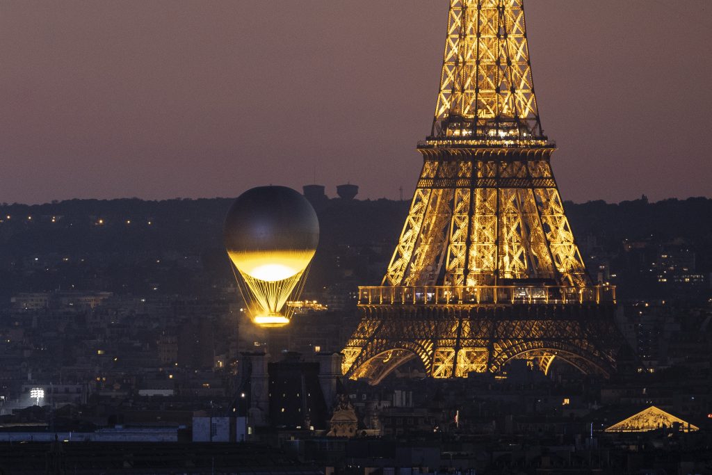 Mathieu Lehanneur's Olympic Cauldron floating in the skies of Paris next to the Eiffel Tower on day fifteen of the 2024 Olympic Games. The top of the illuminated tower is cropped out, with the cauldron, a floating orb that looks like a hot air balloon, illuminated from below, hovering near the base.