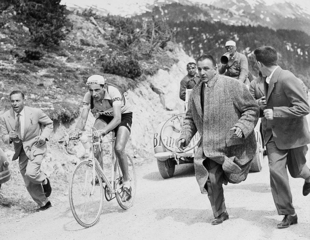 A black and white photo of cyclists at the Olympics with reporters running alongside them.