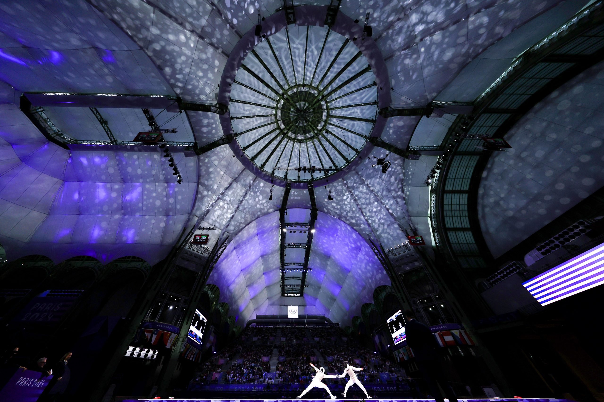 two figures in white can be seen fencing against a vast, spectacular backdrop o fa building with a domed glass roof that is lit up purple