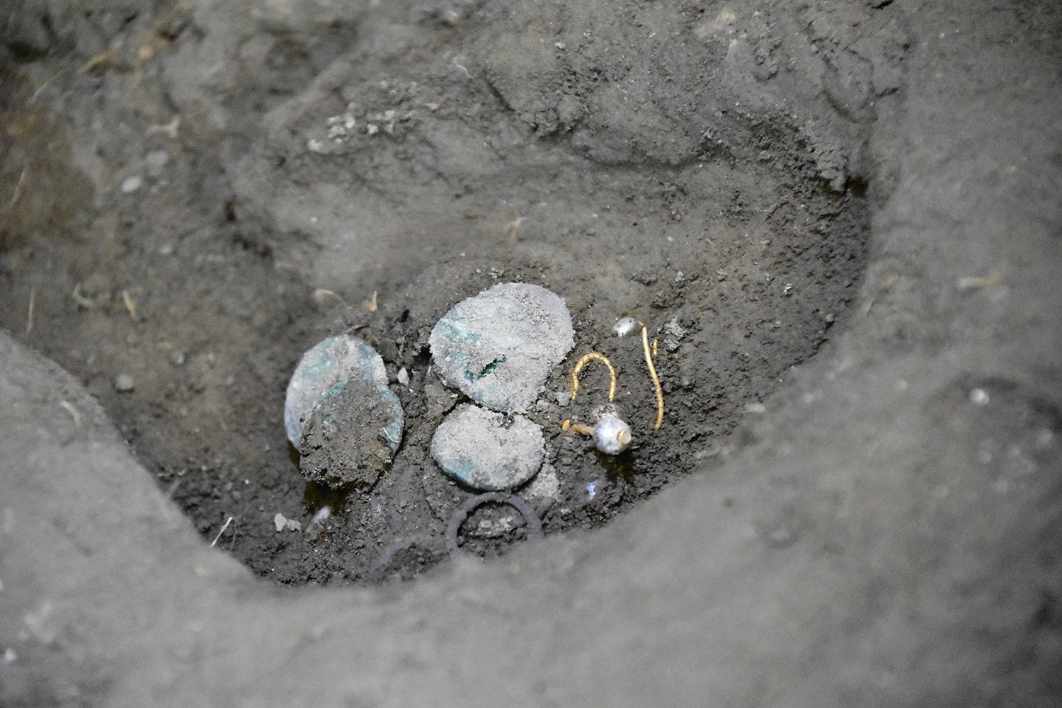 A photograph of ancient coins and pearl earrings half-buried in the dirt of the Room 33 archaeological site at Pompeii