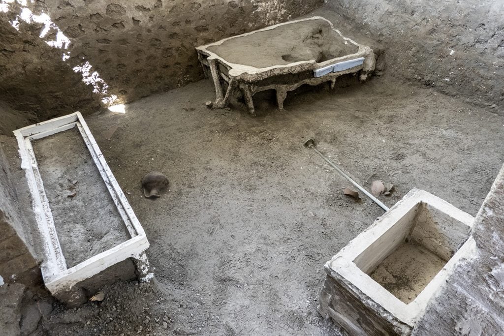 A photograph of the archaeological site of Room 33 at Pompeii—a rectuangular dirt hole populated with three rudimentary pieces of furniture