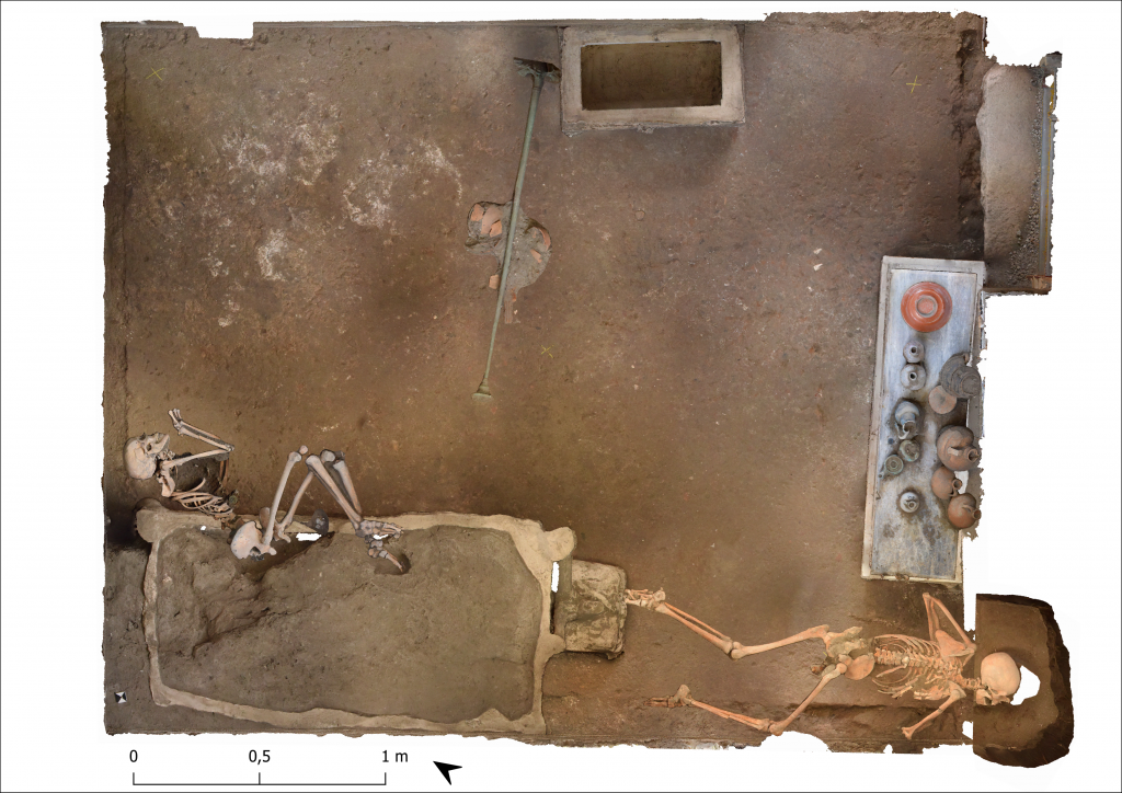 An aerial view of the archaeological site of Room 33, with a skeleton in the fetal position in the room's lower left corner, a prone skeleton in the lower right, a table scatter with objects along the right wall, and another rectangular piece of furniture along the top wall