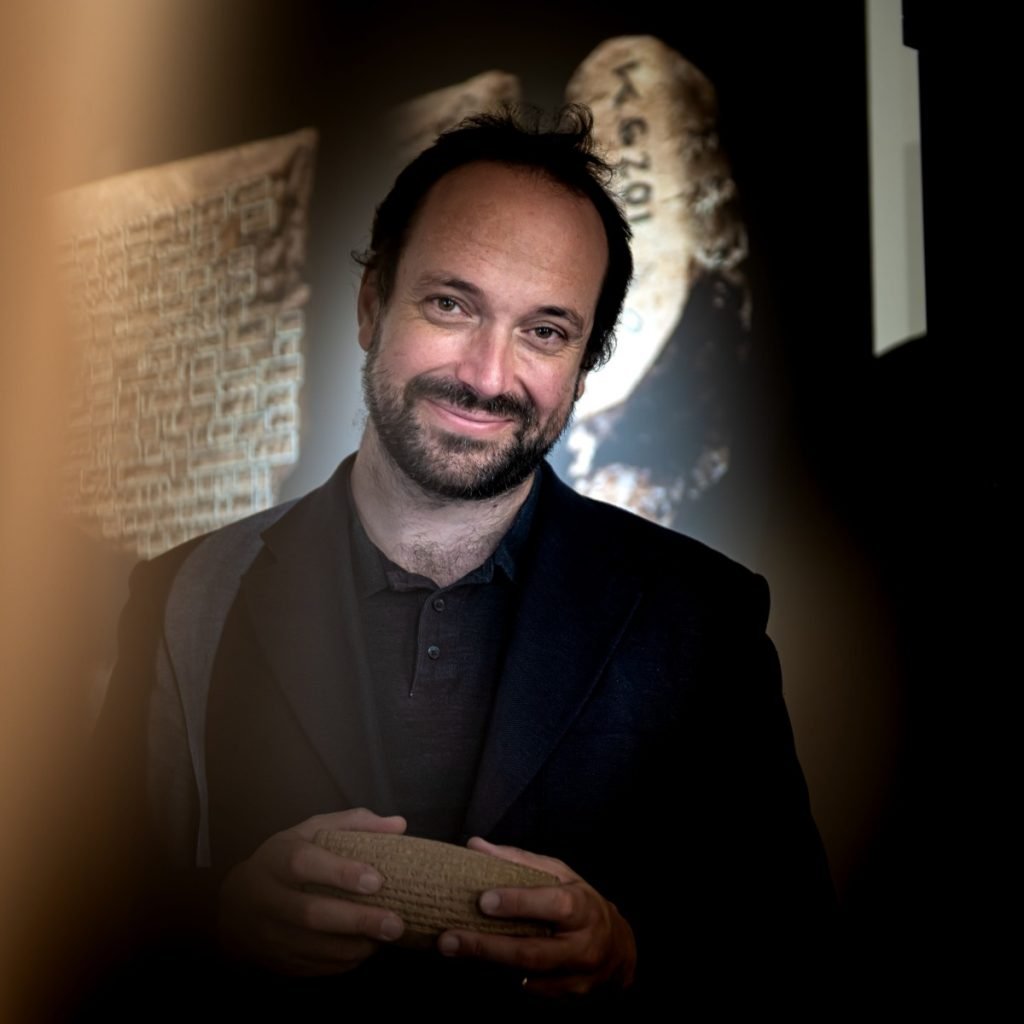 Institute of Assyriology professor Enrique Jiménez, a man with dark hair and a short beard in a black shirt and jacket, stands in front of large photos of cuneiform tablets in a darkened room.