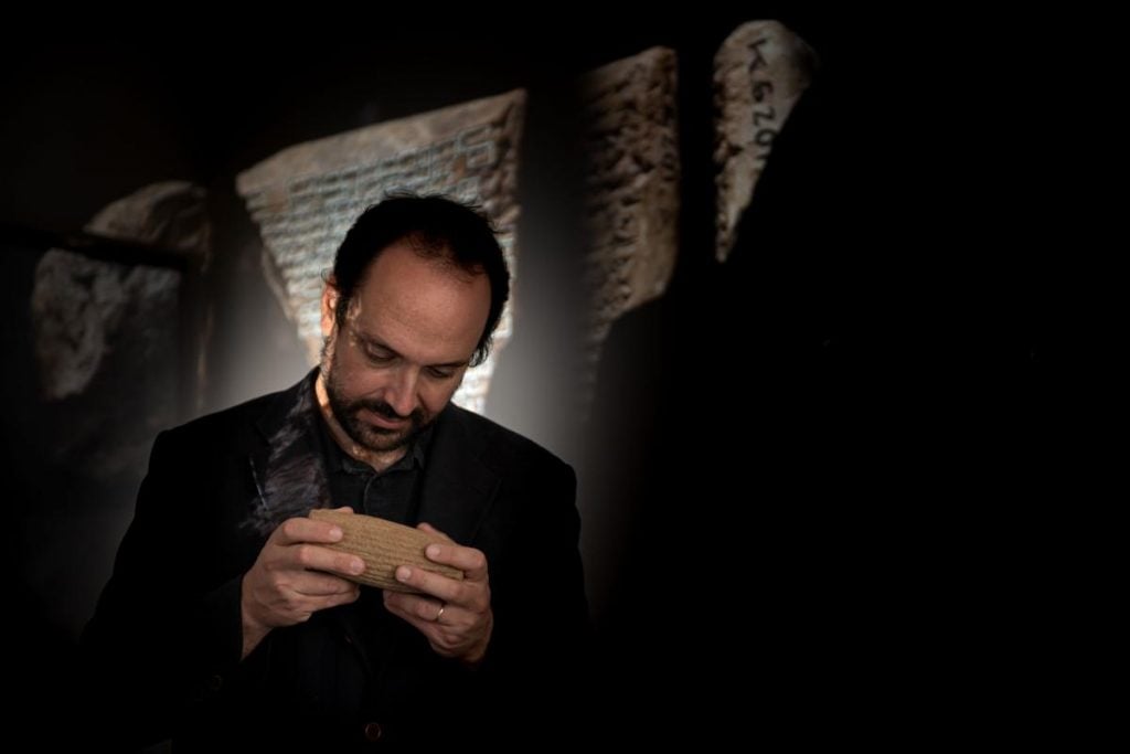 Institute of Assyriology professor Enrique Jiménez, a man with dark hair and a short beard in a black shirt and jacket, stands in front of large photos of cuneiform tablets in a darkened room. He is looking down at an artifact in his hands. 