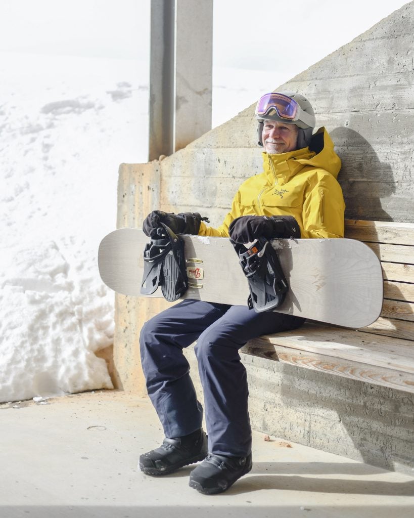 A man in winter clothing with a snowboard poses for the camera.