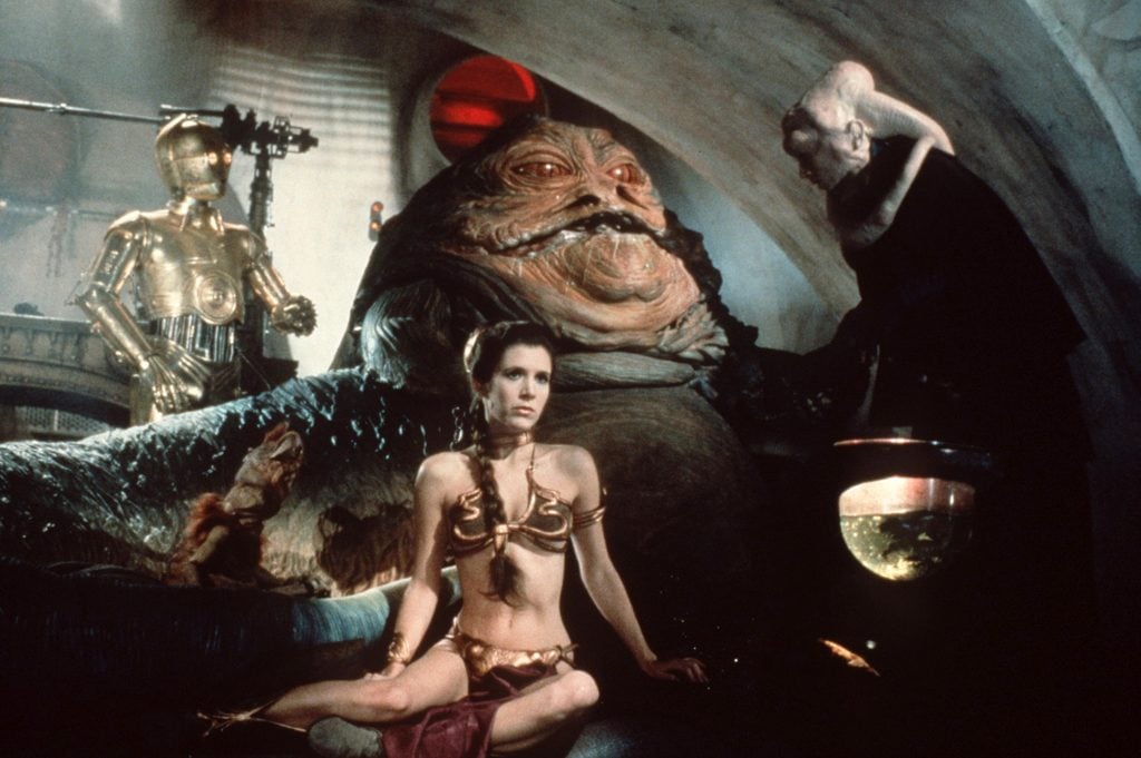 A still from Star Wars: Return of the Jedi showing a woman in a gold bikini sitting in front of a giant slug. A robot stands in the background, while an alien with a white tentacled head stands to the side