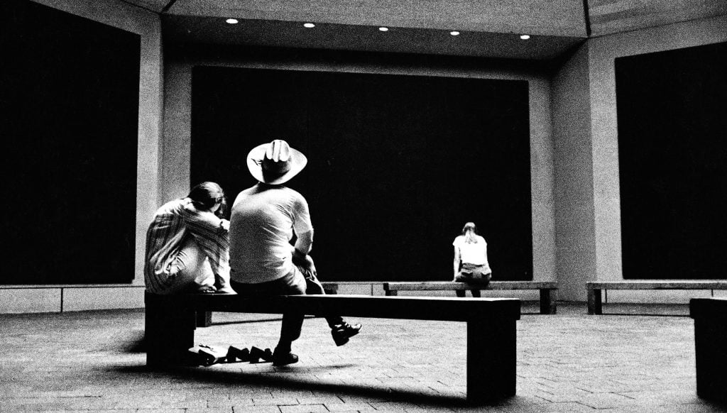 Two men and a woman sitting on benches in the Rothko Chapel, an octagonal room hung with huge black paintings