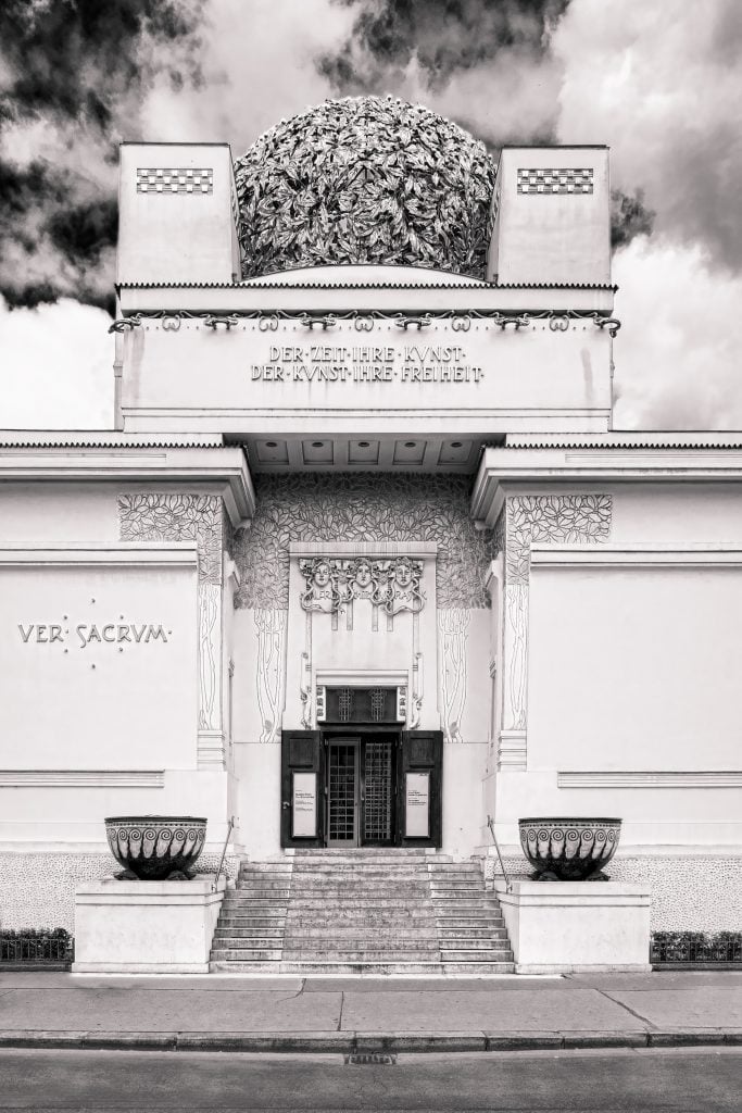 Black and white photograph of the Secession Building in Vienna, showing a grand white facade with gold letters