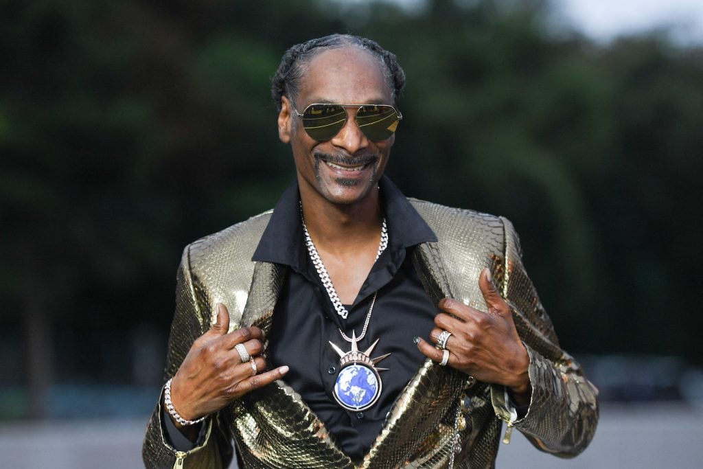 Rapper Snoop Dogg posing in a golden crocodile suit, holding open the lapels of his blazer to reveal a chain with an image of a globe