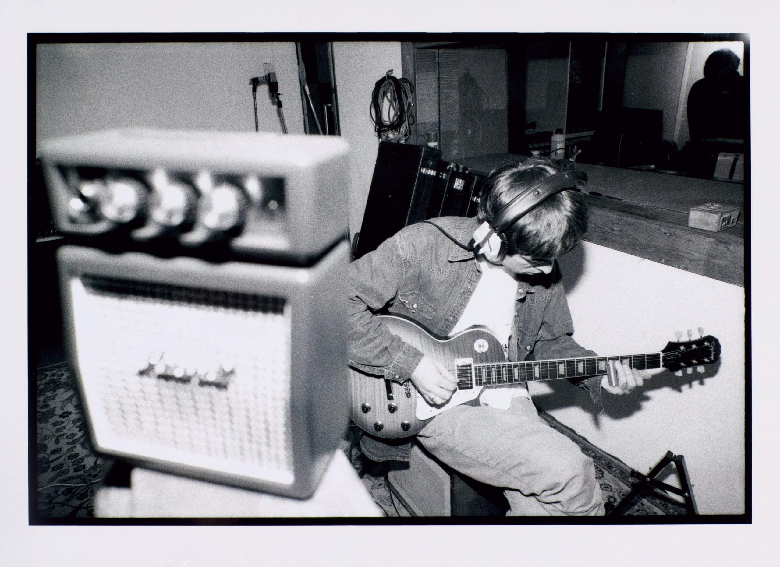 A black and white photograph of Oasis frontman Noel Gallagher seated and playing a guitar, with an amp to his left