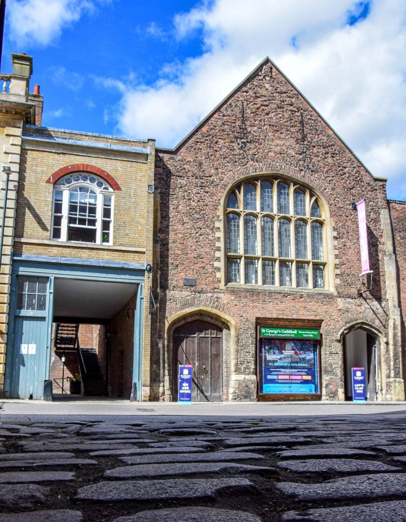 A photograph of a pointed, two-story brick guild house with a large arched window on a cobblestone street under a blue sky