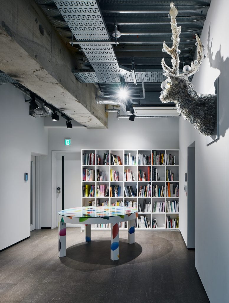 A room with a large bookshelf, an ornate round table and a sculpture of a deer covered in soap bubbles on the right wall.