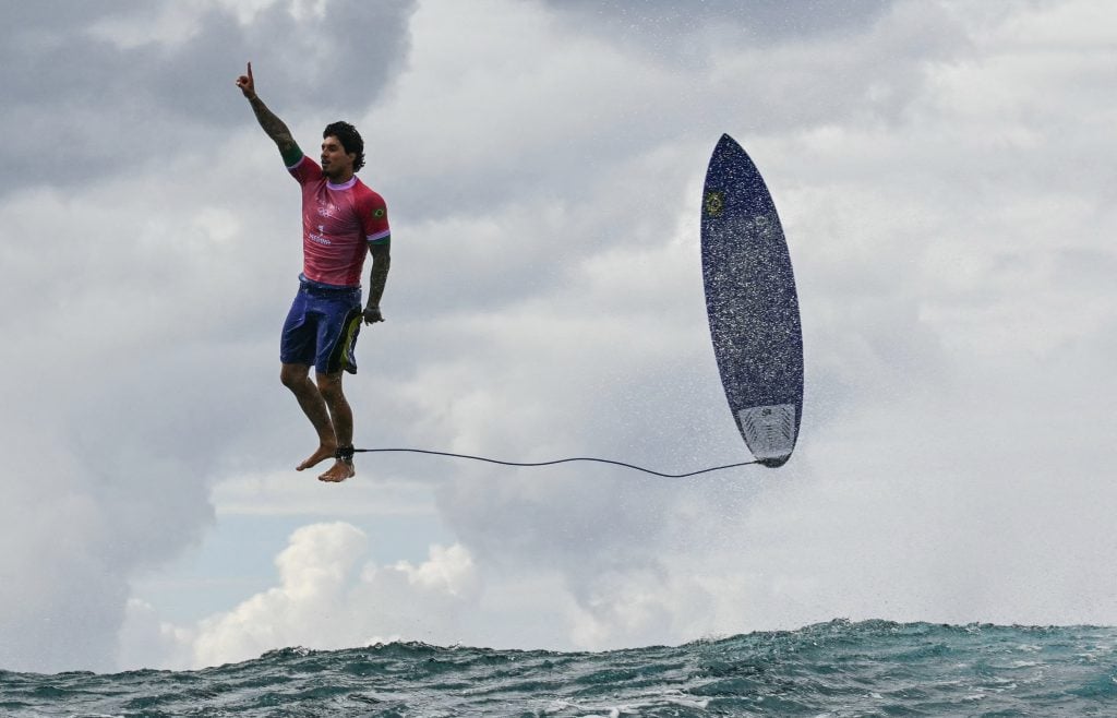 One of the most recognizable photographs from the 2024 Paris Olympic Games was this shot of Brazil's Gabriel Medina reacting after getting a large wave in the men's surfing competition in Teahupo'o, on the French Polynesian Island of Tahiti. A dark haired young man in a red spandex top and blue bathing suit trunks has flown off his board, which is suspended in the air parallel to him above the surf, in front of a cloudy sky. He points up in triumph.