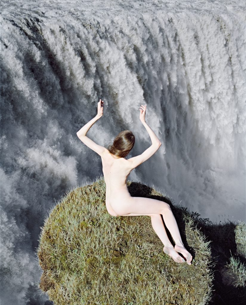 A woman nude on a grassy ledge above a massive waterfall with her arms upstretched.