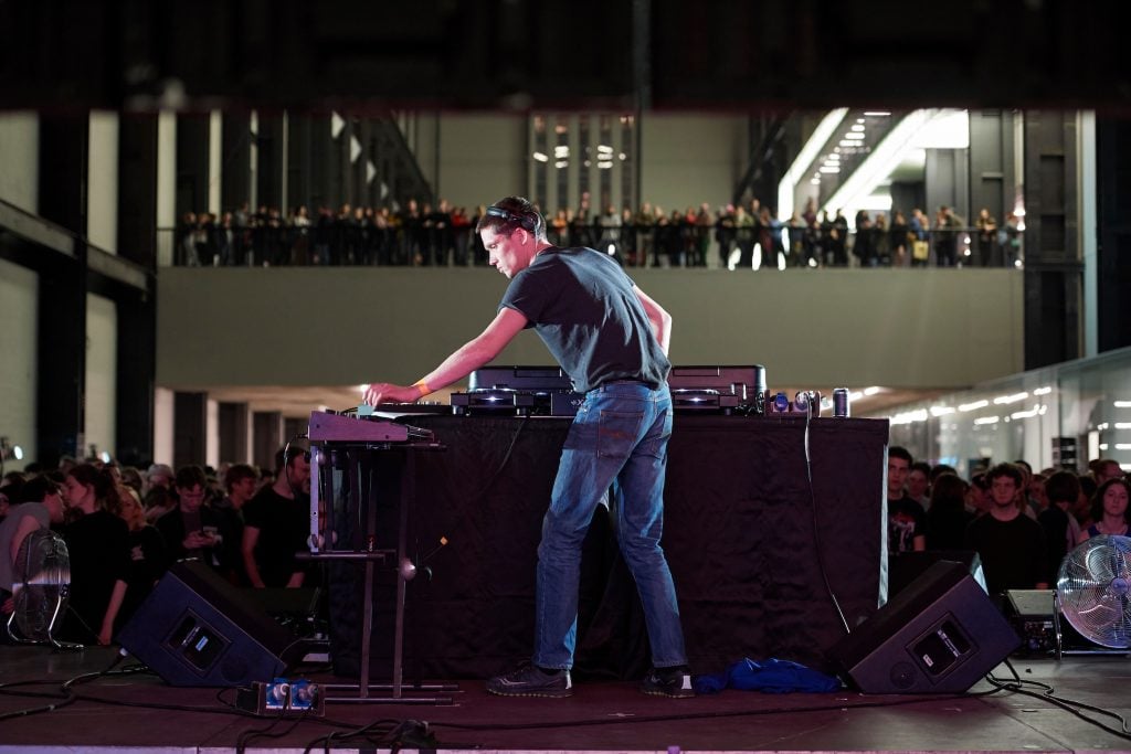 A photograph of a DJ spinning records before two stories of dancers in a large, dark, sparse space