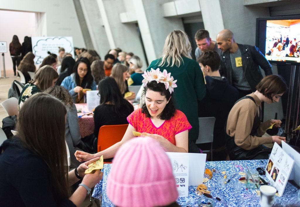 A photograph of a girl at a table crowded with art supplies in a brightly lit room