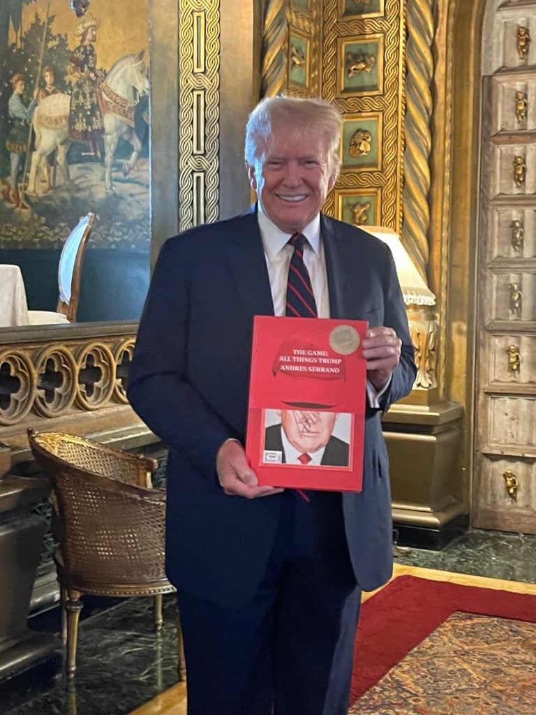 A man in a suit and tie stands in a gold-adorned interior holding a red catalogue that says "THE GAME: ALL THINGS TRUMP"