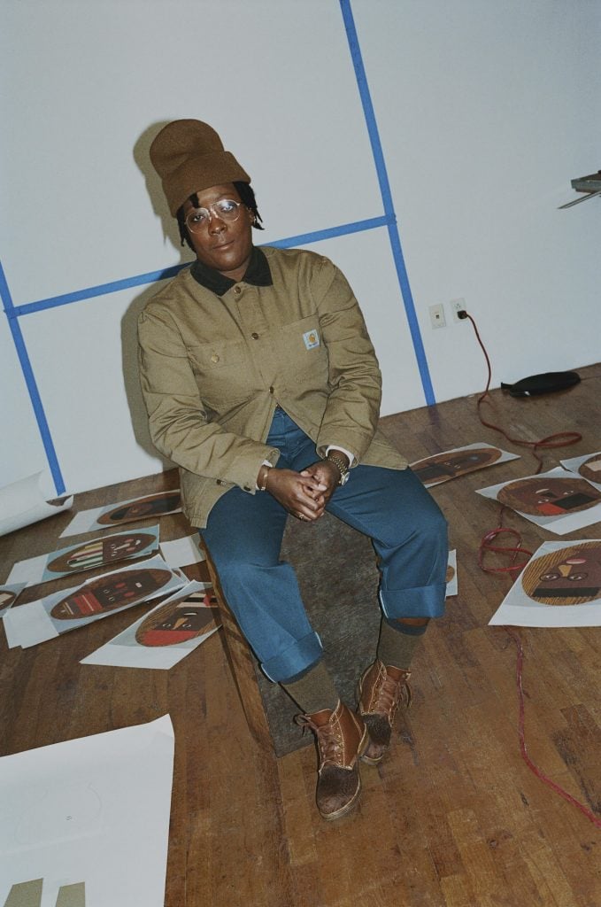 a black woman wearing a brown beanie, a hunting jacket, jeans and ll bean rainboot sits on a chair. some artworks are visible scattered on the floor around her.