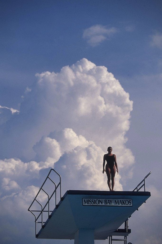 A diver at the Olympics at a high dive platform silhouetted against a large formation of white fluffy clouds and blue sky.