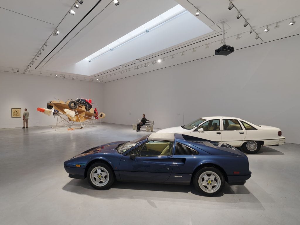 an industrial space filled with one blue car and one white car, plus a deconstructed car sculpture