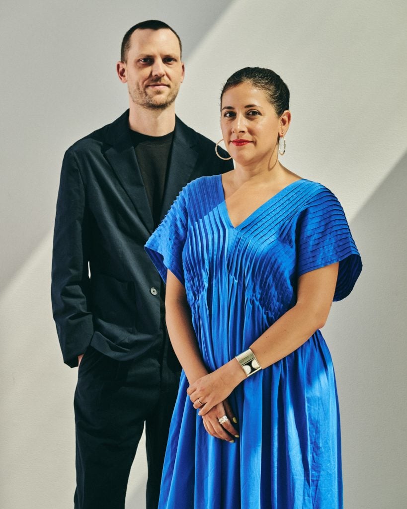 A photograph of Drew Sawyer and Marcela Guerrero, the 2026 Whitney Biennial curators at the Whitney Museum of American Art in New York. He is a young white man with short dark hair and a short beard wearing all black with a suit jacket over a t-shirt. She is a young Latino woman wearing red lipstick, a pleated bright blue dress with large sleeves, and hoop earrings, with her hair pulled back. They are photographed down to just below the waist, in front of a white wall with shadows on it.