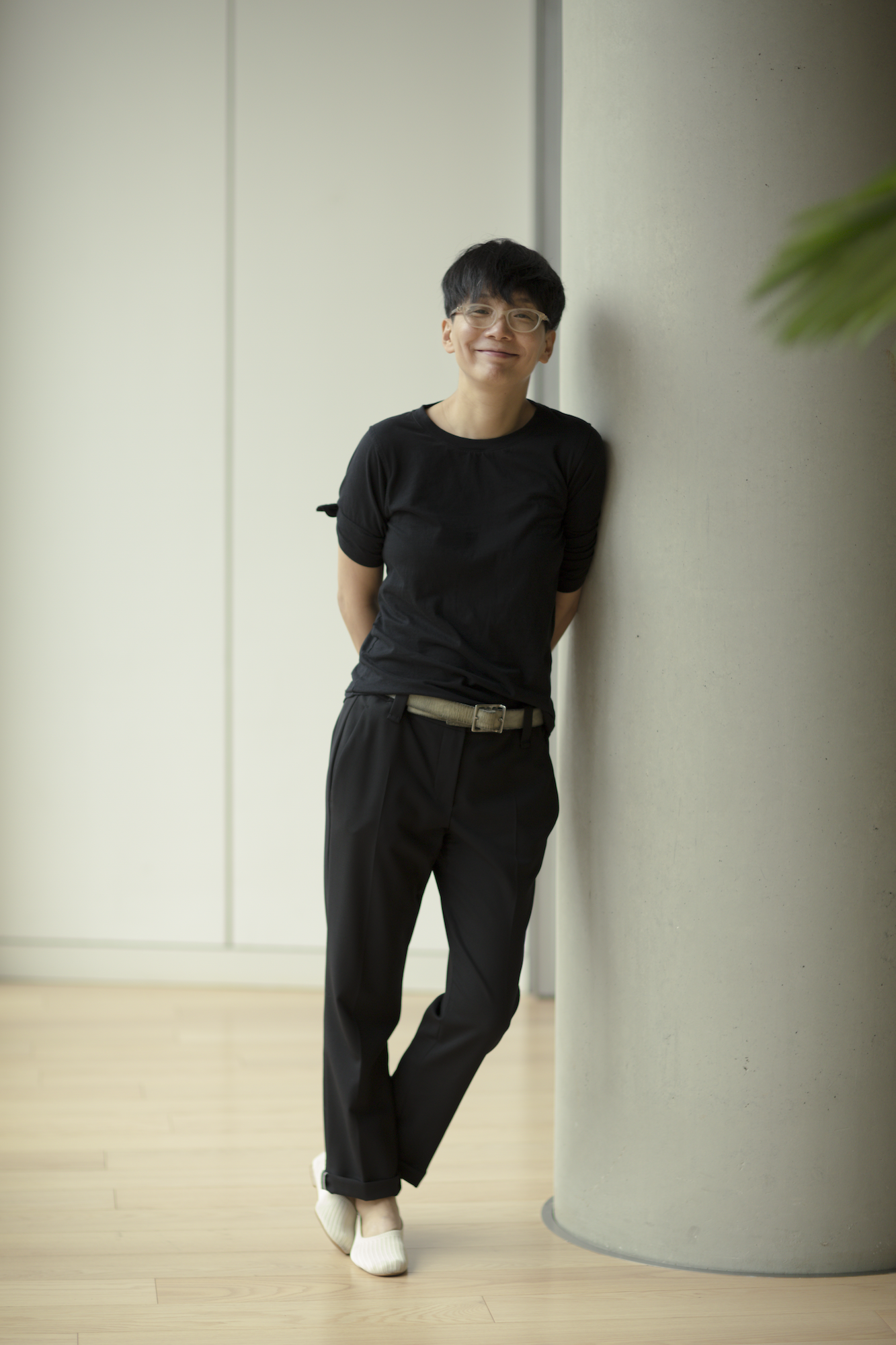 a portrait of an asian person with short black hair and glasses, leaning on a column, and wearing a black teeshirt and black pants