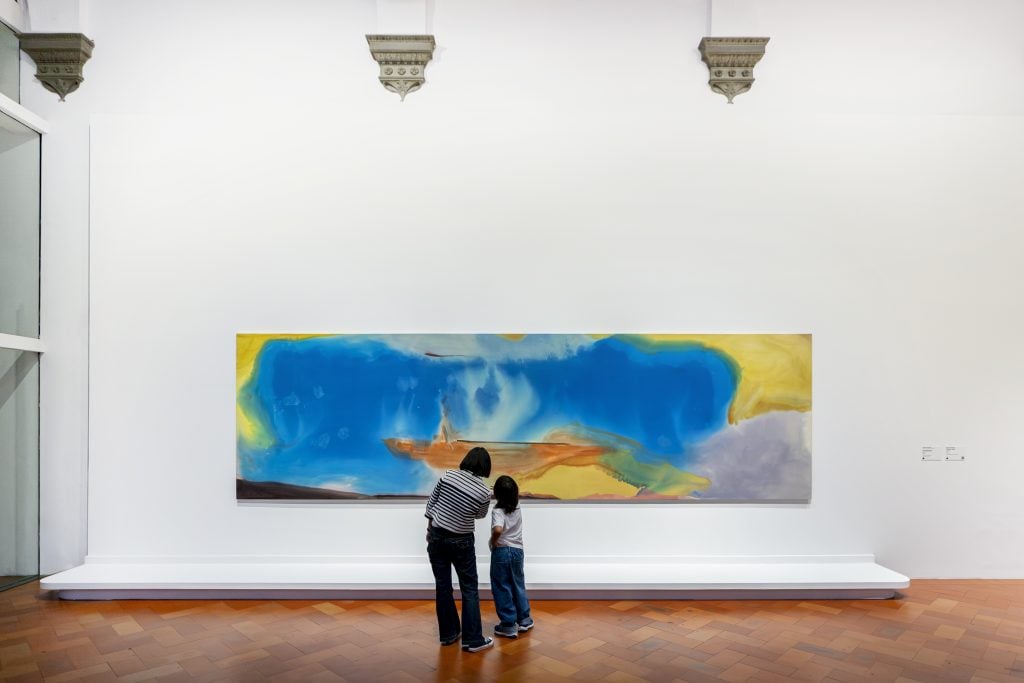 People standing in front of a large blue and yellow painting by Helen Frankenthaler 