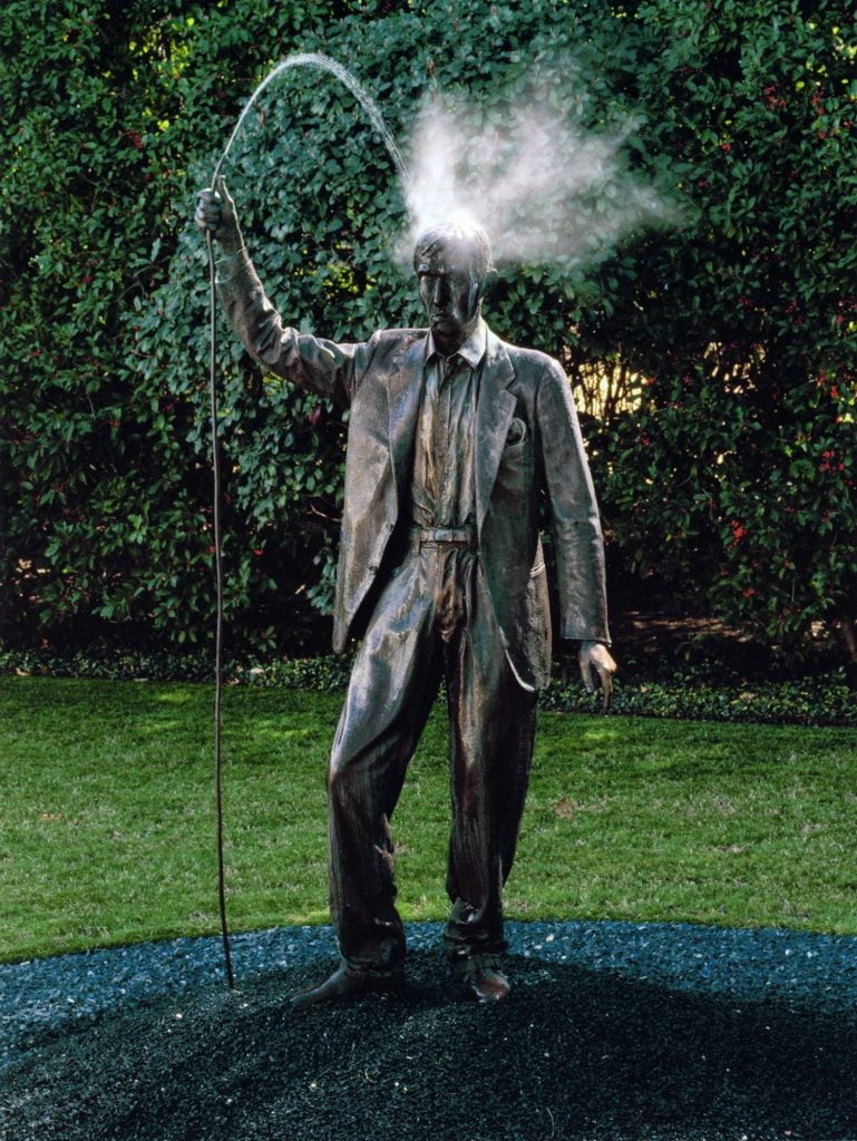 The image shows a bronze statue of a man in a suit holding a rod, from which water sprays out and arcs over his head. The figure, seemingly caught mid-action, has water misting dramatically from the top of his head. The statue stands on a patch of dark ground, with greenery in the background, creating a striking contrast between the natural surroundings and the artificial, dynamic fountain-like effect of the sculpture. This artwork is Autoritratto (Self-portrait) by Alighiero Boetti, combining both a sculptural presence and functional water fountain elements.