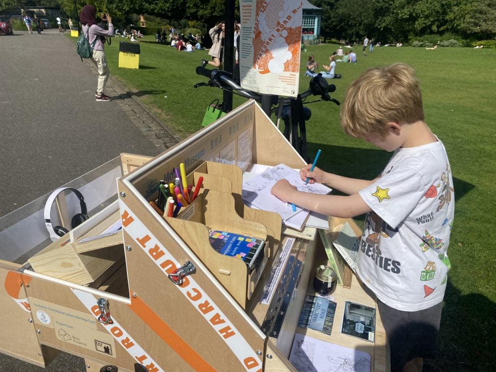 A blonde hair child colouring a piece of white paper on on shelf, in front of green grassland