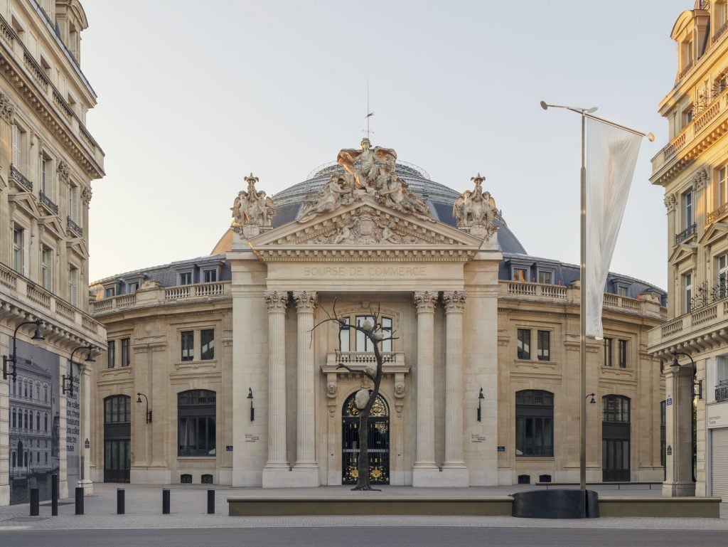 A wide view of the Bourse de Commerce building in Paris. The building’s grand neoclassical facade features columns, intricate sculptures, and a dome. In front, there is an art installation featuring a barren tree branch with large rocks on its limbs.