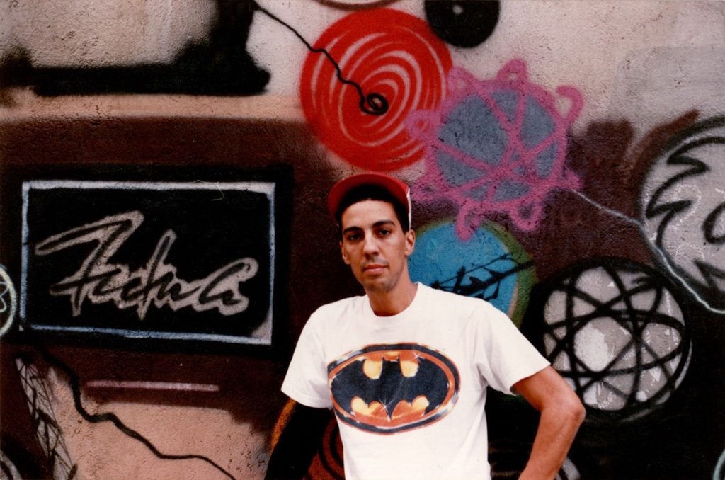 Futura as a young man in Barcelona. The mixed race artist, wearing a red brimmed hat and a white t-shirt with a large Batman logo on it, stands in front of spray painted mural bearing his name.