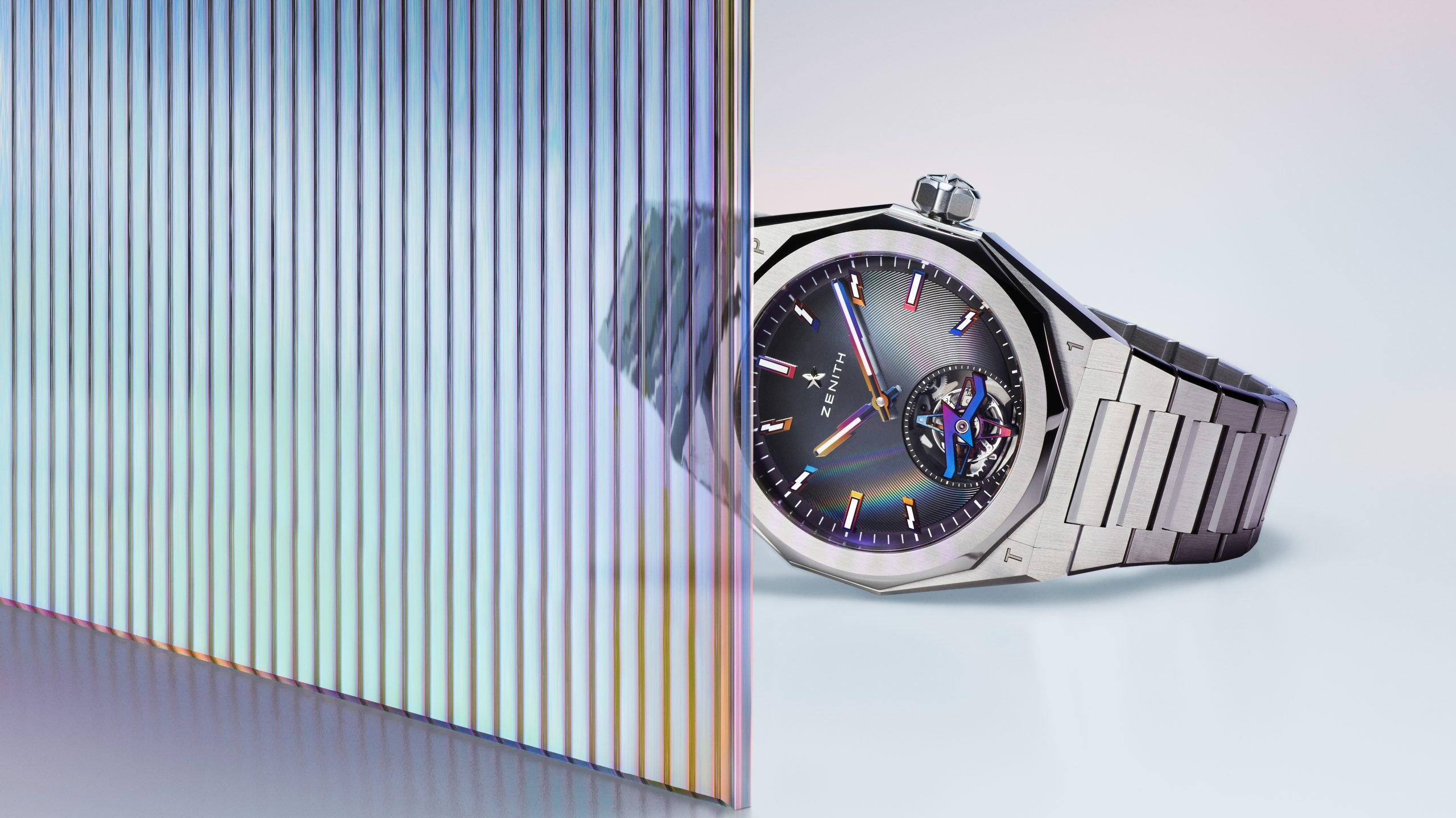 A sleek metallic watch lays on its side behind an irridescent transparent ridged barrier