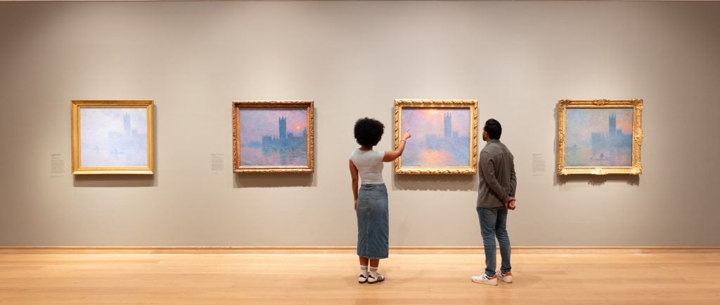 a photograph in which two figures stand facing a series of pink and purple hazy cityscape paintings hanging on the wall inside what is clearly a museum space