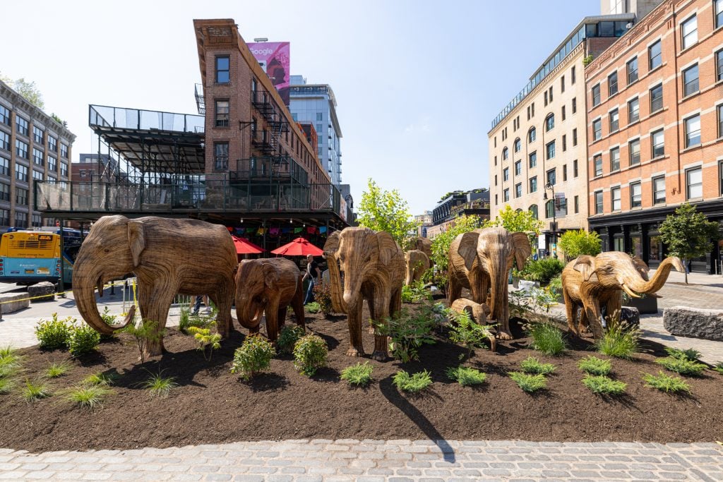 "The Great Elephant Migration" in New York's Meatpacking District. A photograph of a herd of life-size wooden elephant sculptures amid greenery in front of buildings in the city.